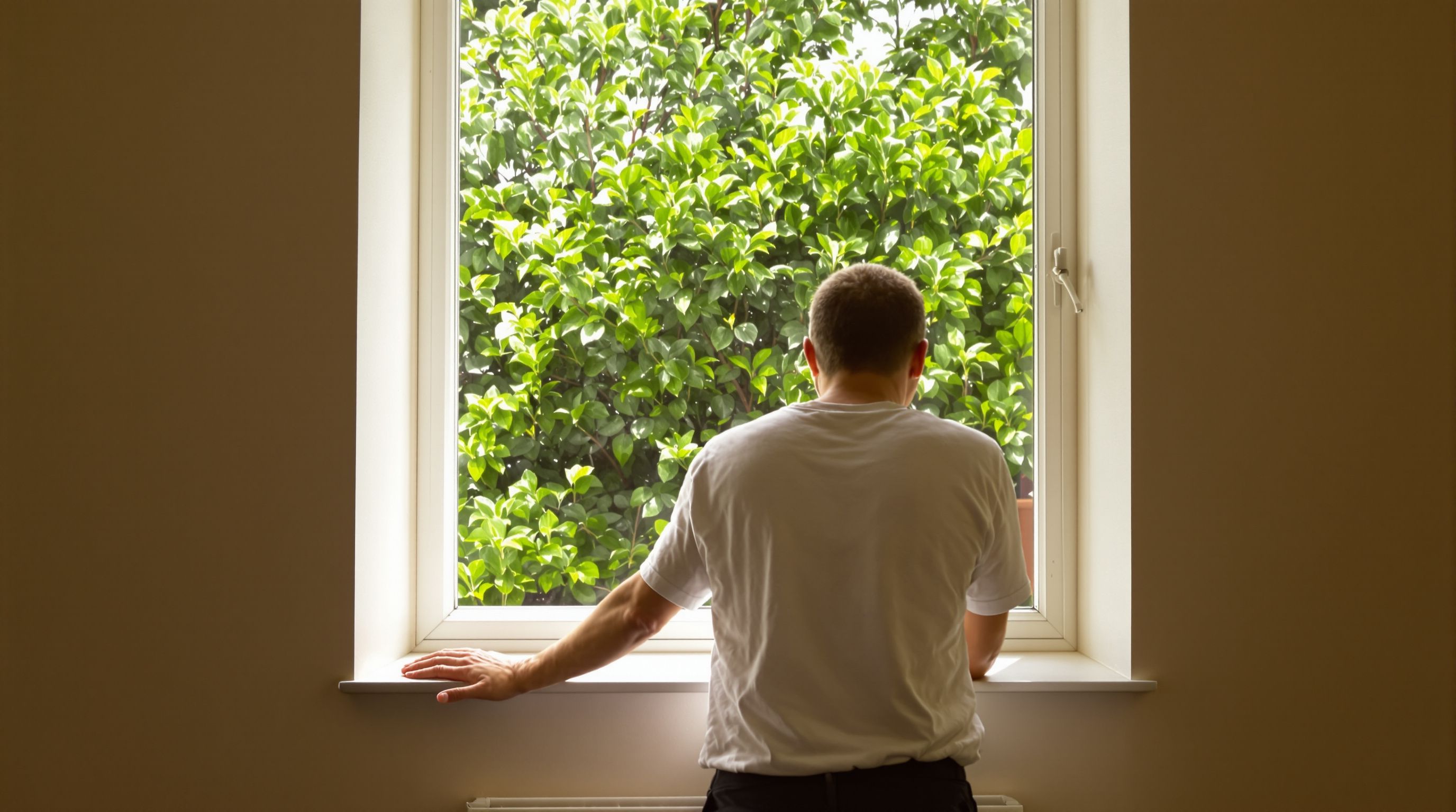 A person standing at a bright window seen from behind, one hand resting on the sill, looking out at green foliage