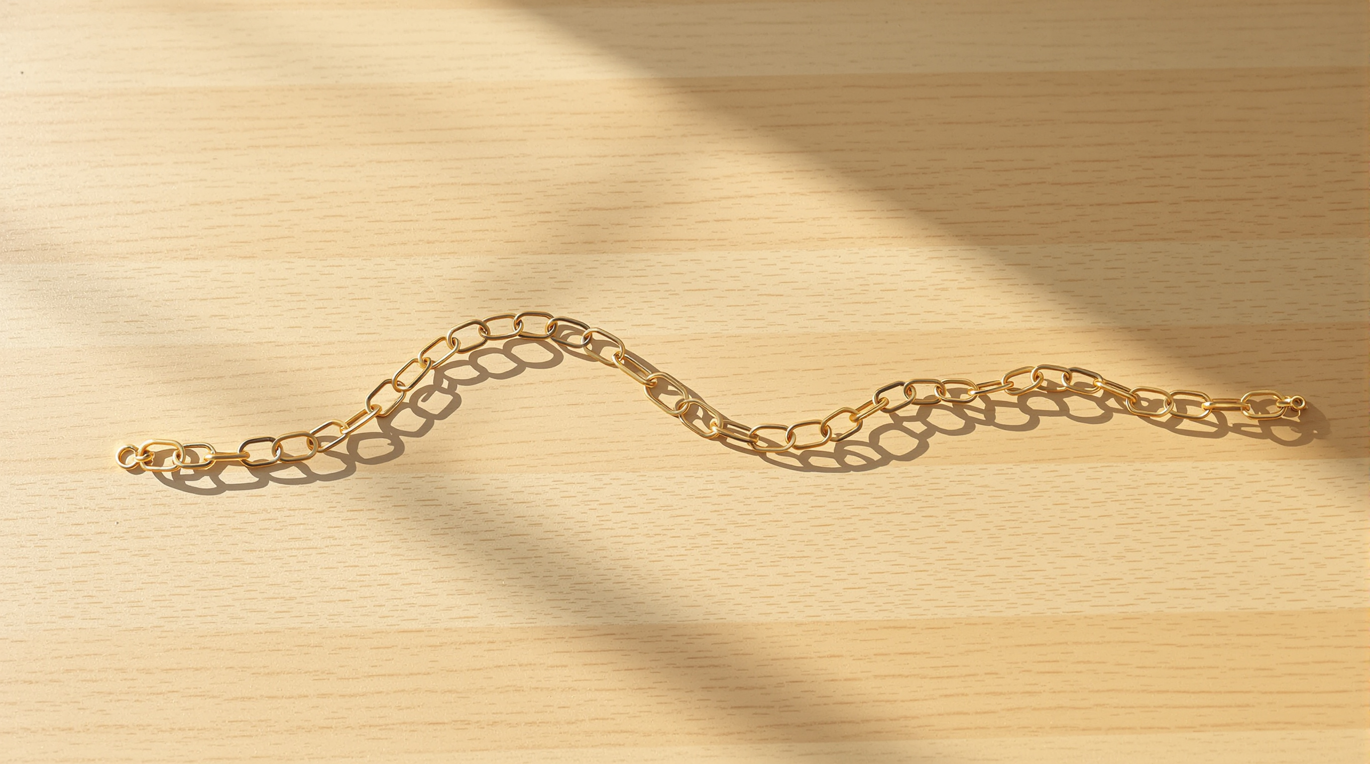 A fine brass chain lying on a pale wooden table in a single curve that rises, dips low in the middle and rises again, in warm morning sun
