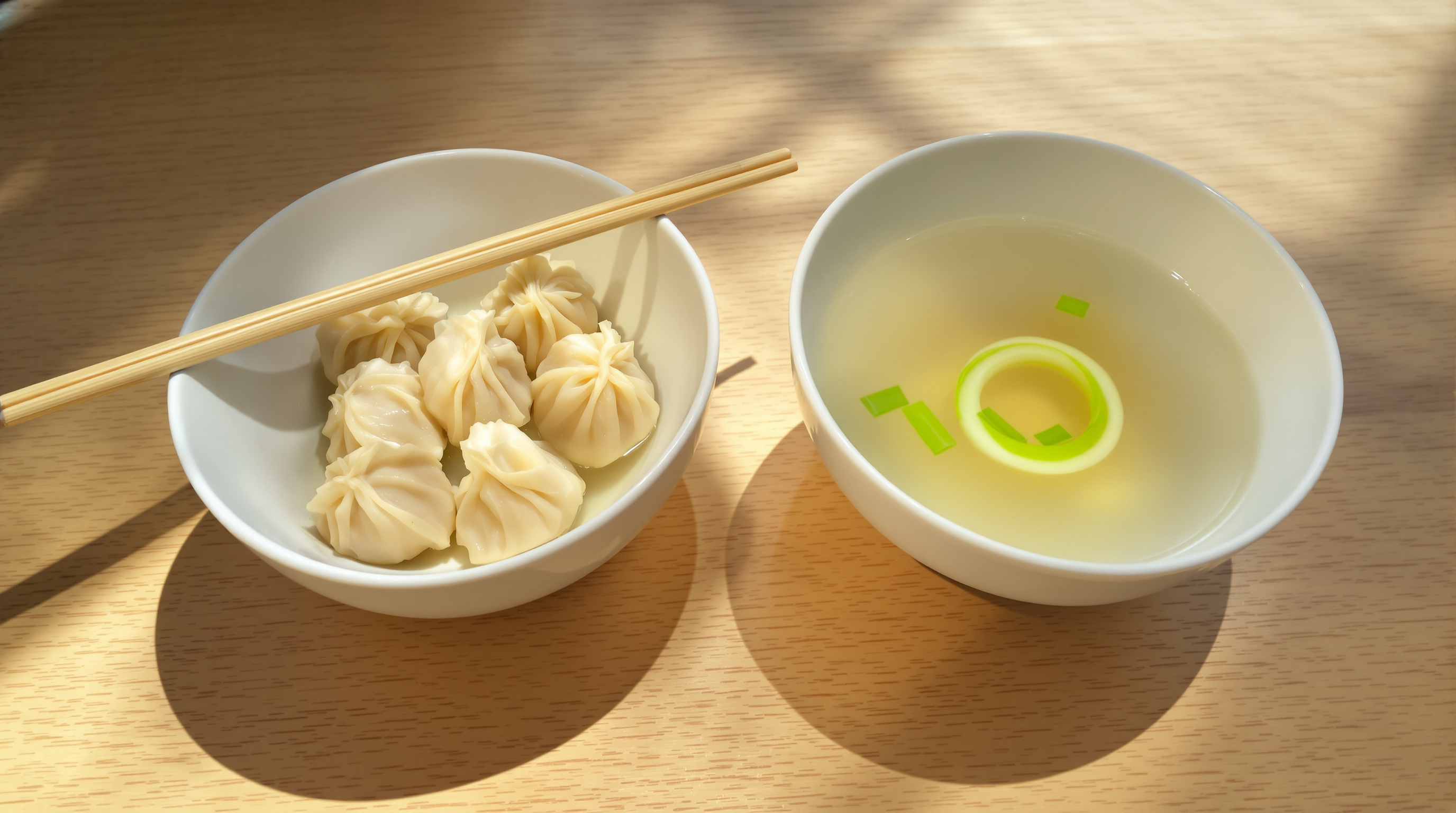 Two small white bowls side by side on a pale wooden table in warm window light, one holding dumplings and one holding clear broth