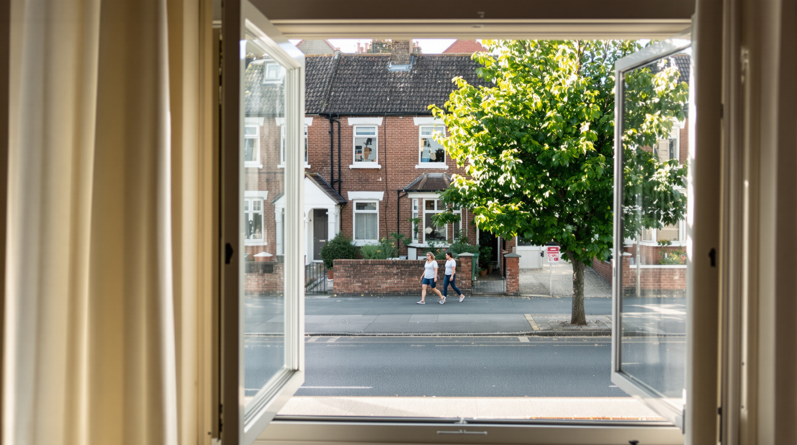 An open window onto a quiet residential street seen from inside a room, two people walking past on the far pavement in late morning sun